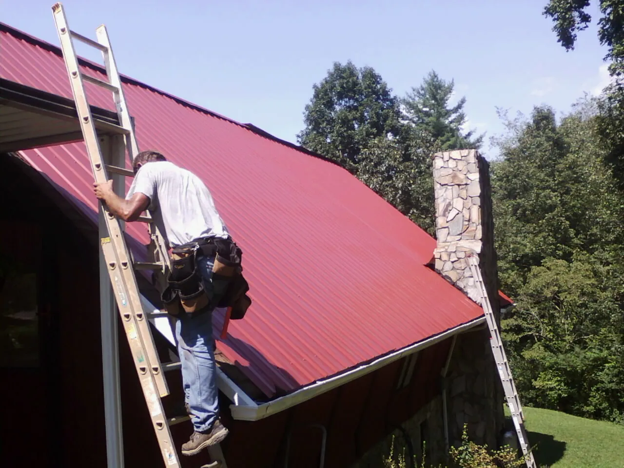 Roofer on a ladder beside a red metal roof and stone chimney — Soffit & Fascia Repair by Roofing Brothers in Kingsland